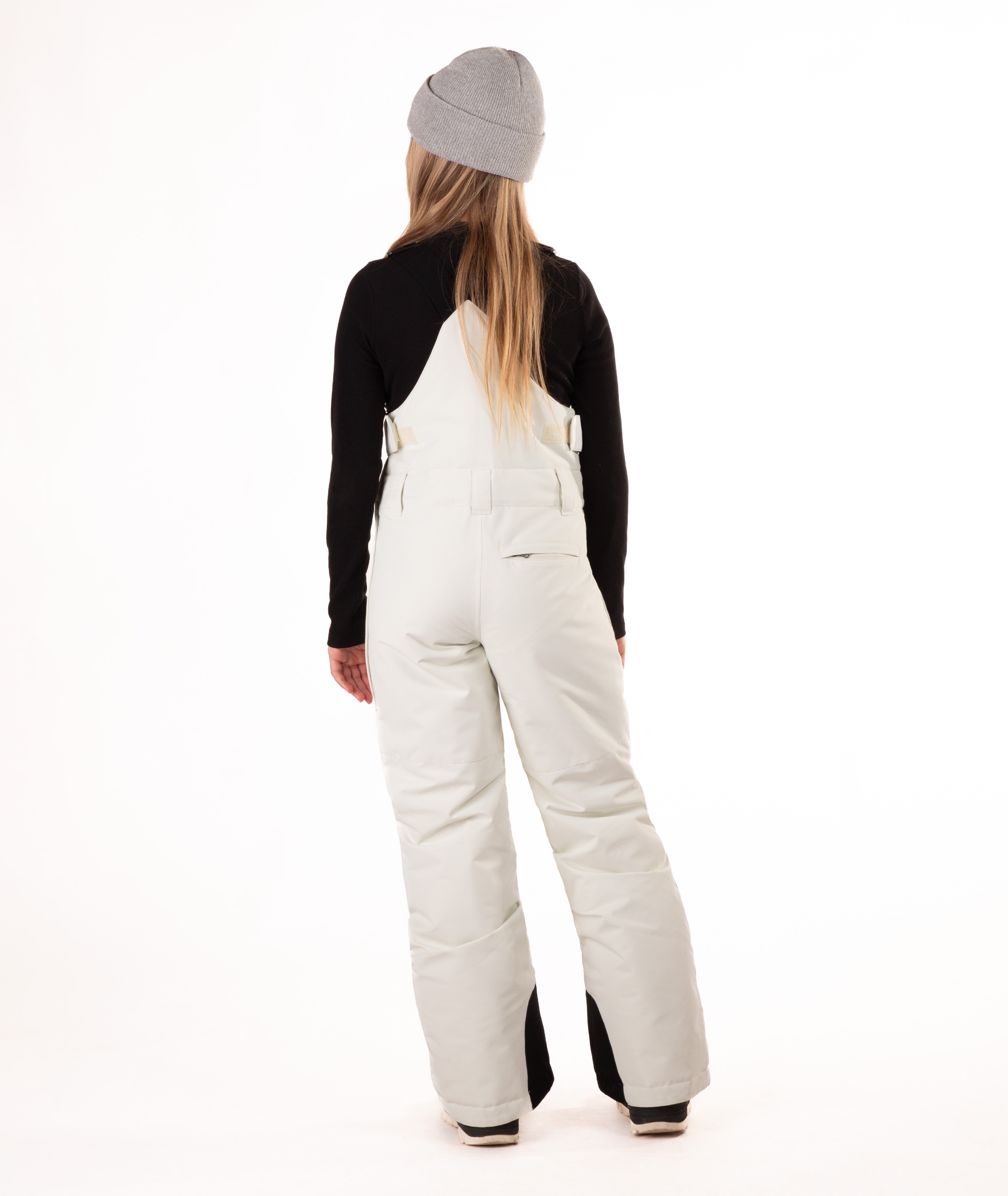 back of girl wearing cream colored snow bibs with a black top on a white background