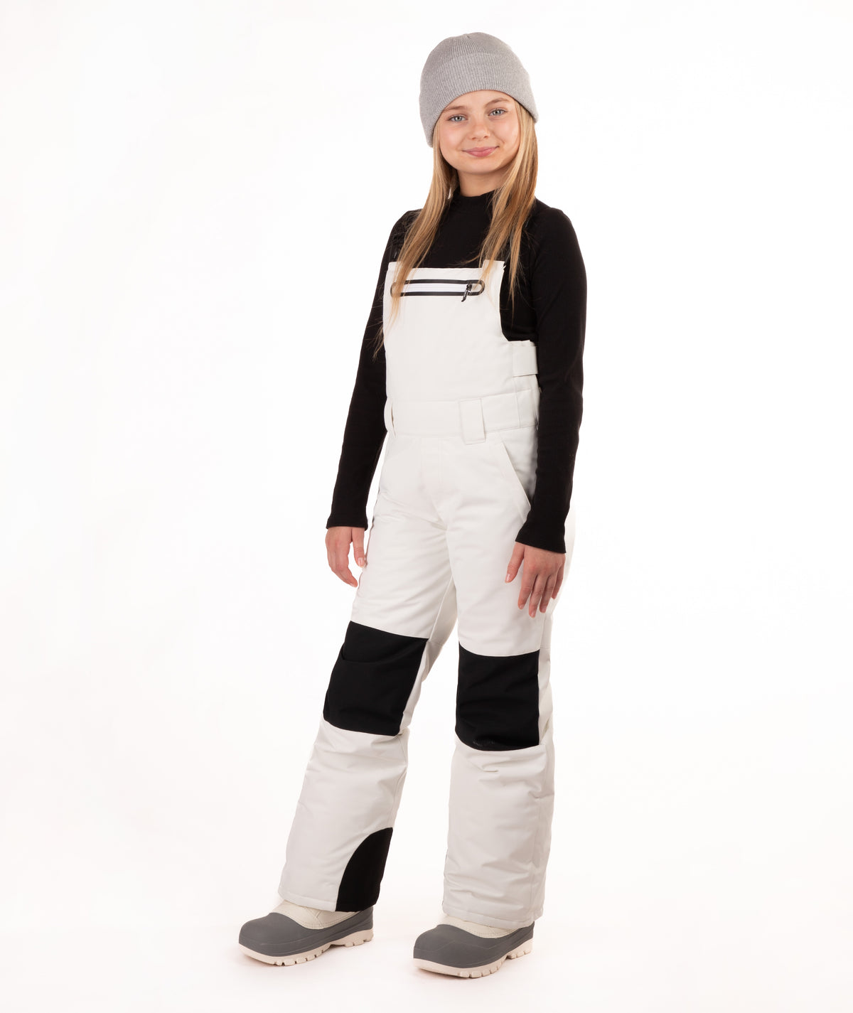 A child model wearing off-white snow bibs with black knee panels, a black long-sleeve shirt, a gray beanie, and gray boots, standing against a plain white studio background.