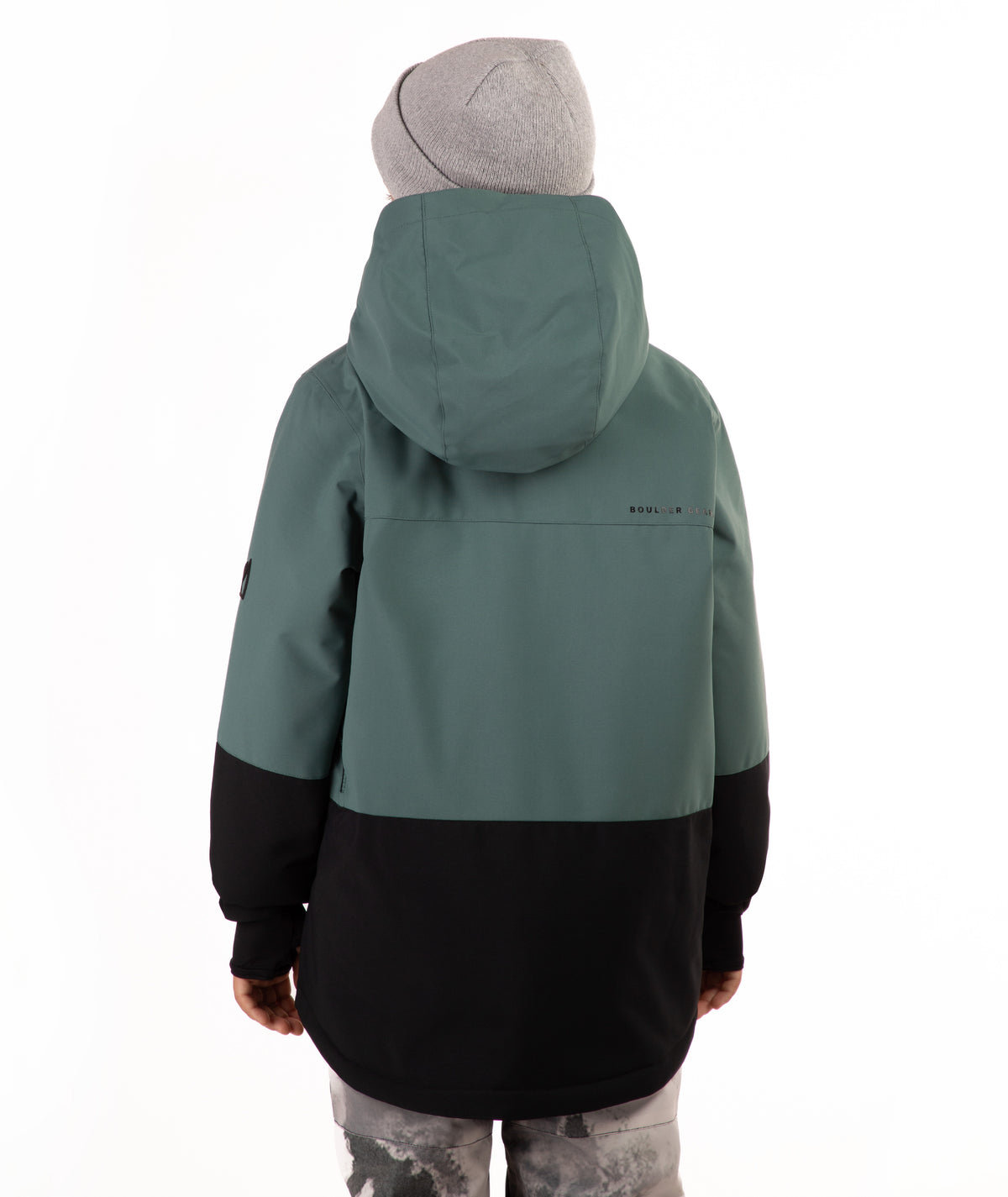 A child wearing a color-blocked ski jacket in sage green and black, paired with a gray beanie and patterned ski pants, standing against a plain white background.