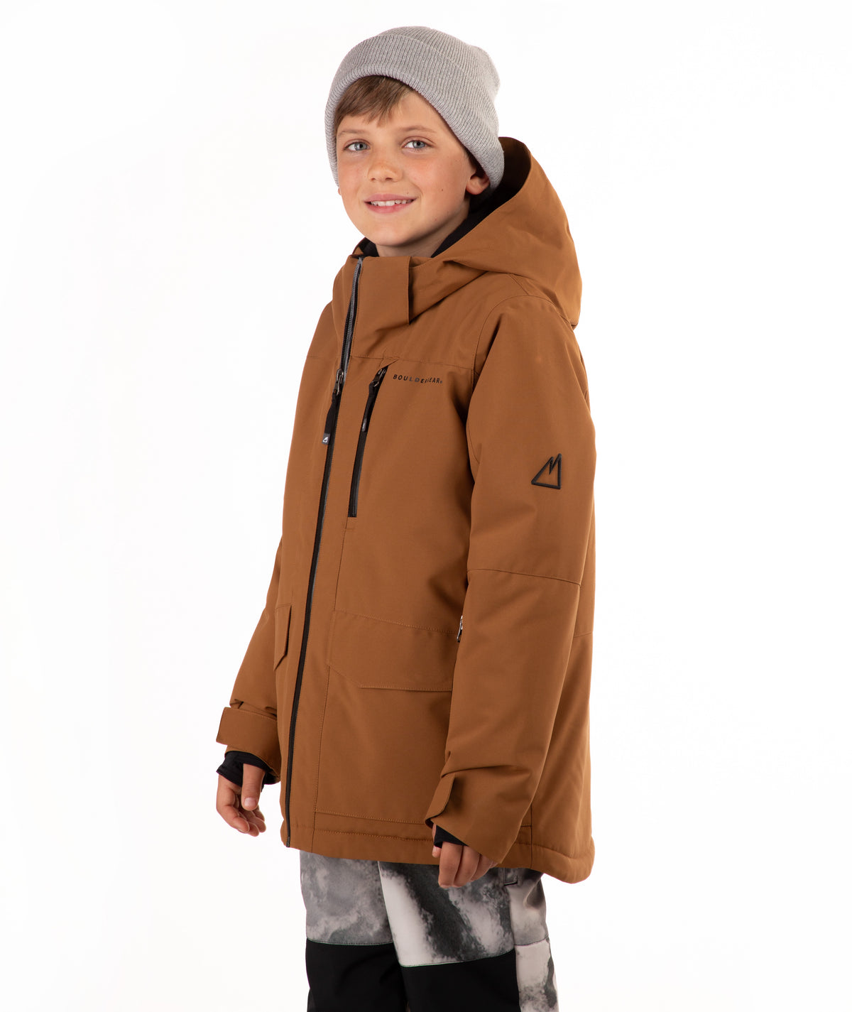 A child wearing a brown hooded snowboard jacket with black zippers, a gray beanie, and patterned pants, standing against a plain white background.