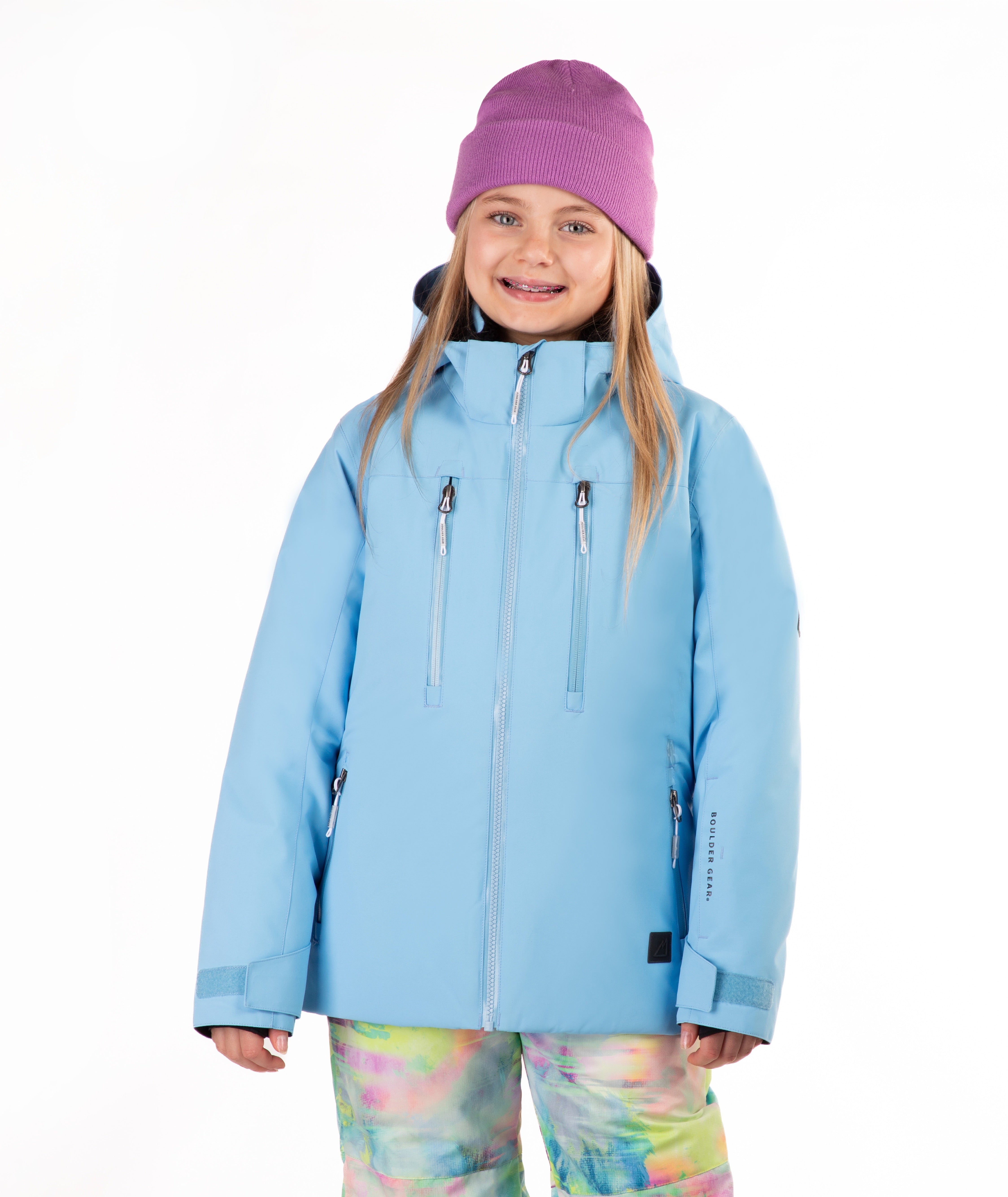 A young girl wearing a light blue ski jacket, a purple knit beanie, and colorful tie-dye snow pants, standing against a solid white background.