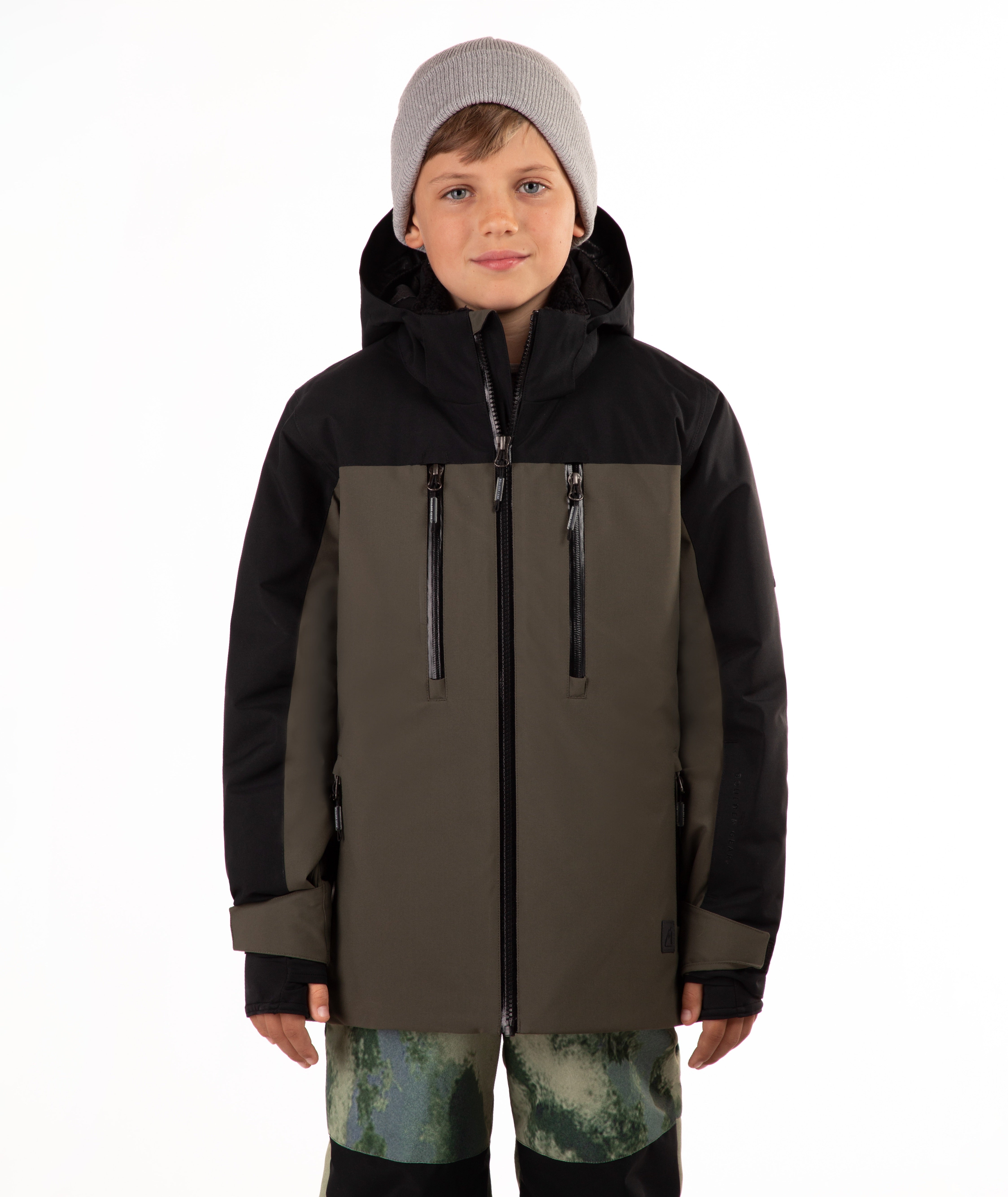 A child wearing a hooded ski jacket in black and olive green, paired with a light gray beanie and green camouflage-patterned pants, photographed against a plain white background.