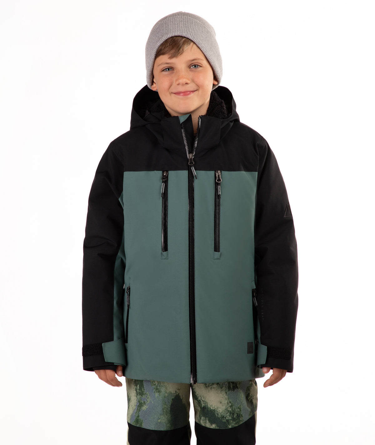 A child wearing a color-blocked ski jacket in black and muted teal, paired with a gray beanie and camouflage-patterned pants, against a plain white background.