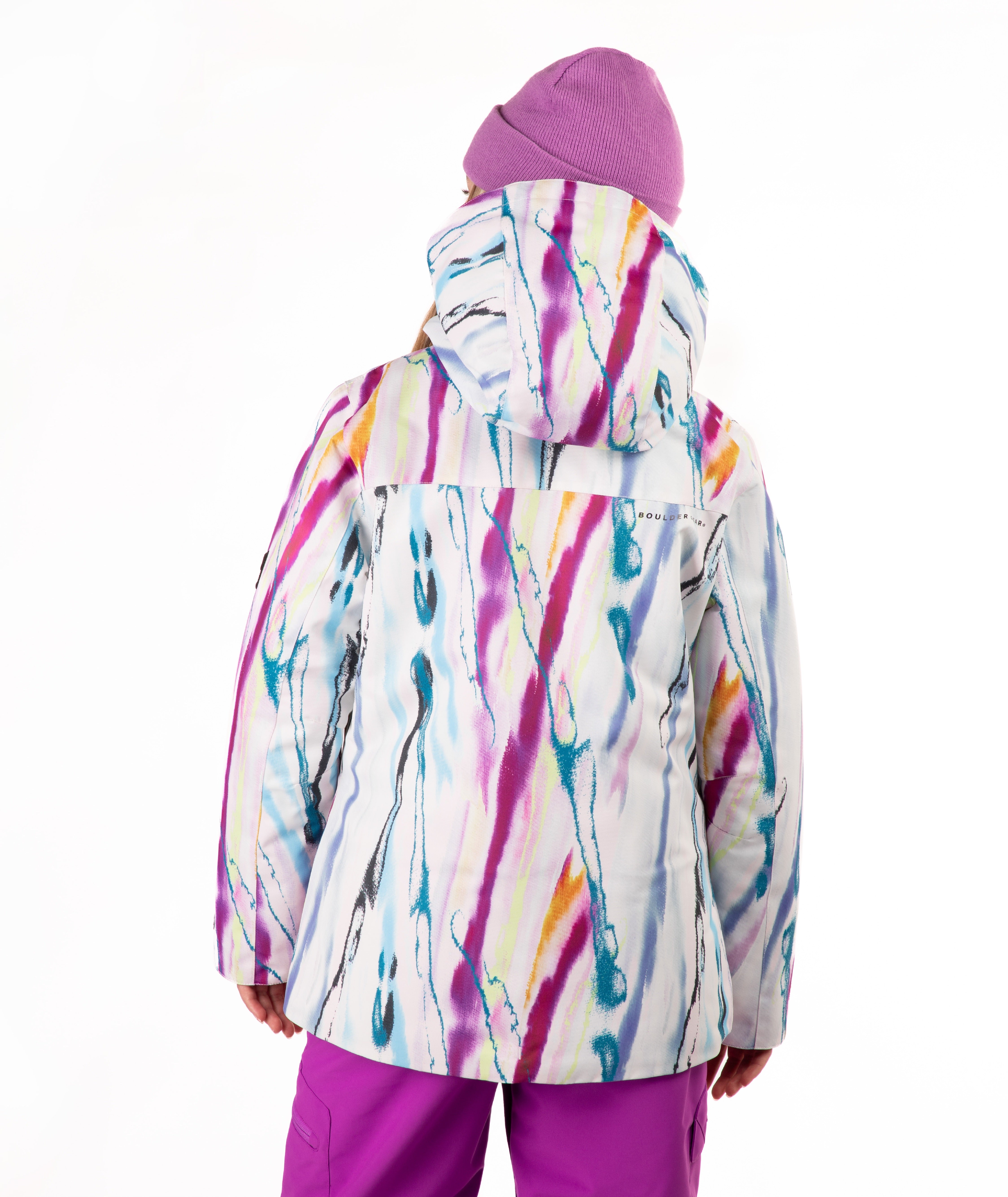 A child viewed from the back wearing a hooded ski jacket with a colorful abstract paint-stroke pattern in pink, purple, blue, yellow, and orange on a white base, paired with a purple beanie and purple pants, against a plain white background.