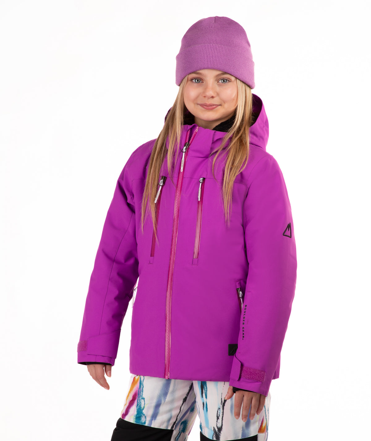 A young girl wearing a bright pin-purple ski jacket, a matching knit beanie, and colorful patterned ski pants, standing against a plain white studio background.