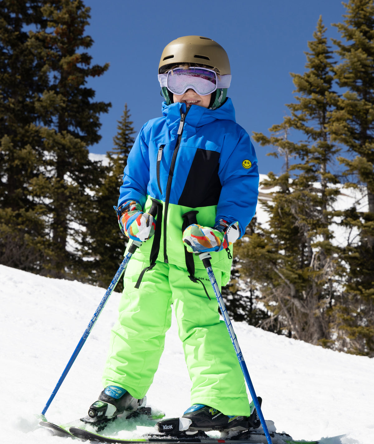 A child skier wearing a bright blue and neon green ski jacket and pants, along with a helmet, goggles, and ski poles, posing on a snowy mountain slope under a clear blue sky.