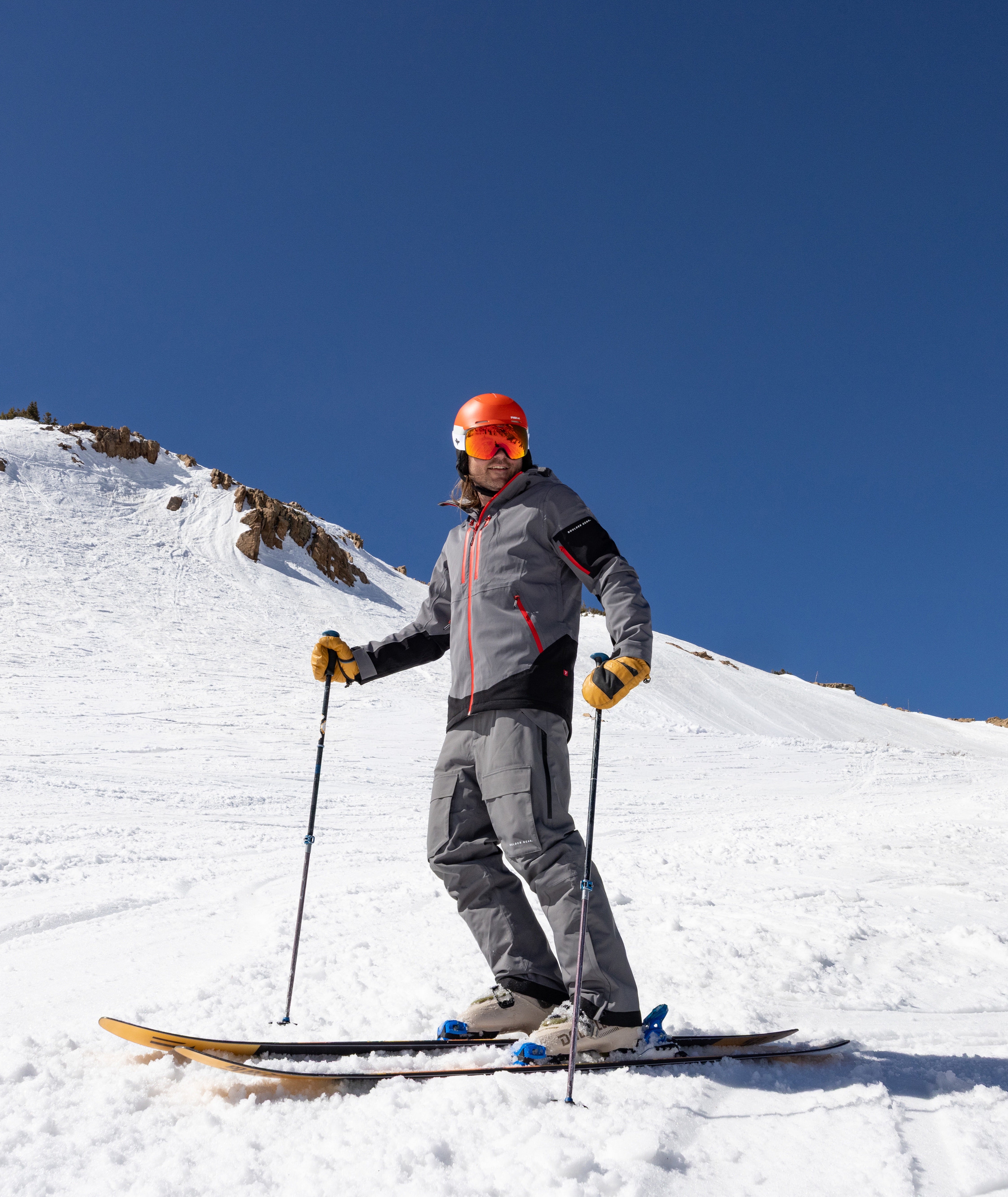 A skier standing on a snowy mountain slope under a clear blue sky, wearing a gray ski jacket and pants with black and red accents, a bright orange helmet, and ski poles.