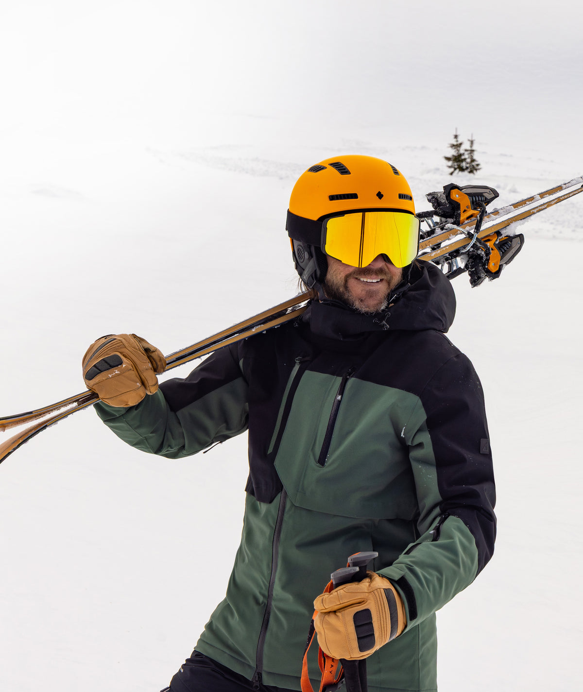 A smiling skier wearing a green and black ski jacket, a bright yellow helmet, and yellow mirrored goggles, carrying ski poles over his shoulder on a snowy mountain.