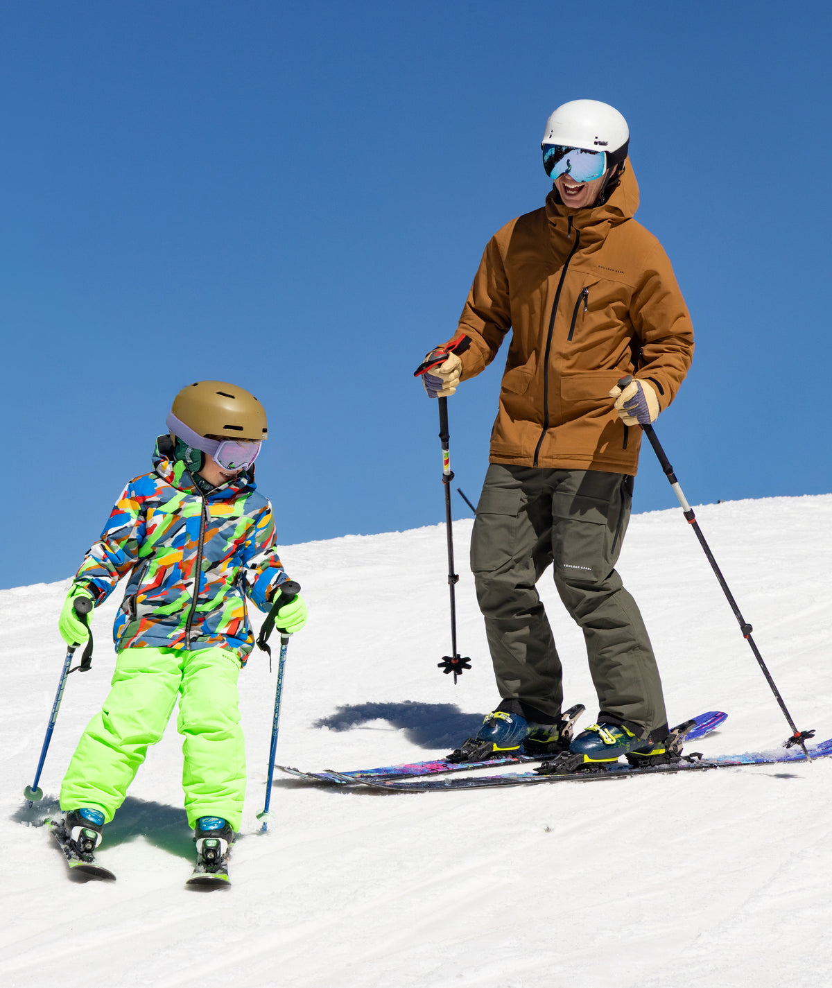 A man and a child skiing on a snowy slope under a clear blue sky. The man wears a brown ski jacket, dark pants, a white helmet, and blue goggles, while the child wears a bright multicolor jacket, neon green pants, and a tan helmet.