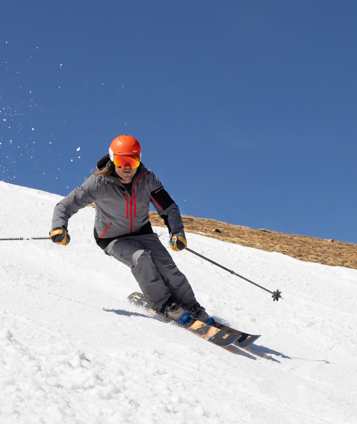 A skier carving through fresh powder on a snowy slope under a clear blue sky, wearing a gray ski jacket, dark pants, a bright orange helmet, and orange-tinted goggles.