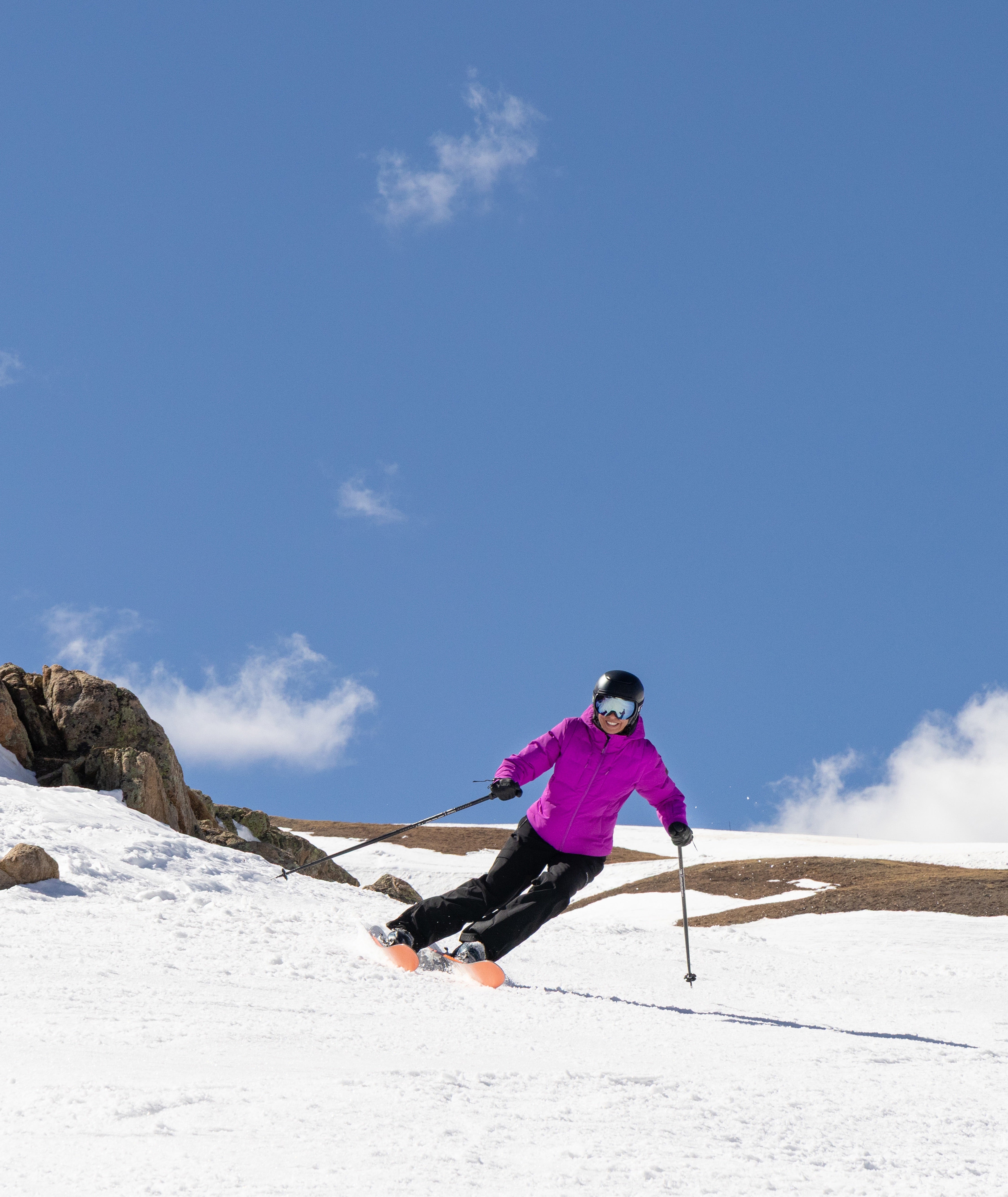 woman shown skiing on a mountain top wearing a bright purple-pink jacket with black pants and helmet.
