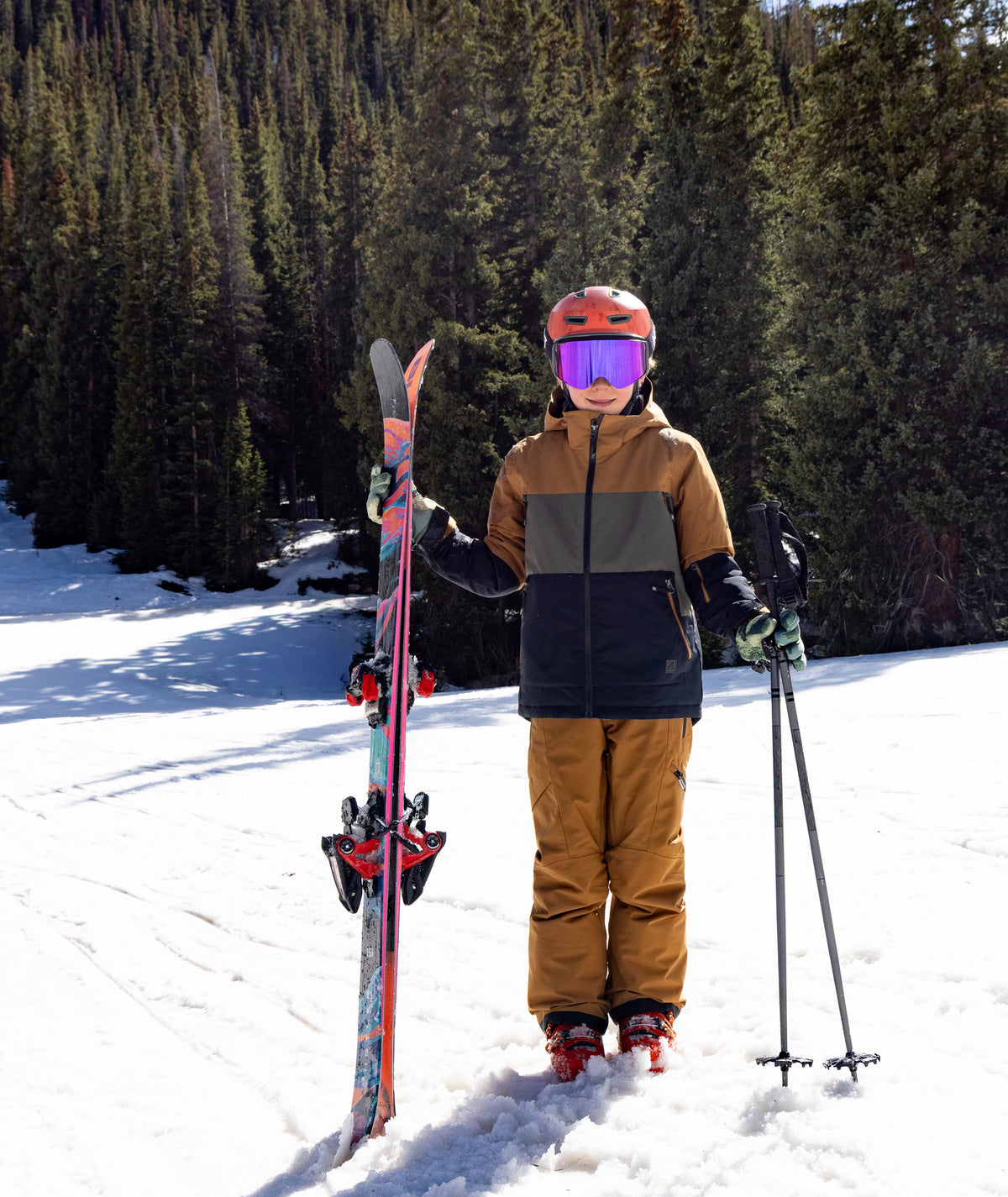 A young person standing in the snow wearing ski gear, including a tan and dark green ski jacket, brown ski pants, an orange helmet, and purple goggles, holding a ski and ski poles with a ski standing vertically.