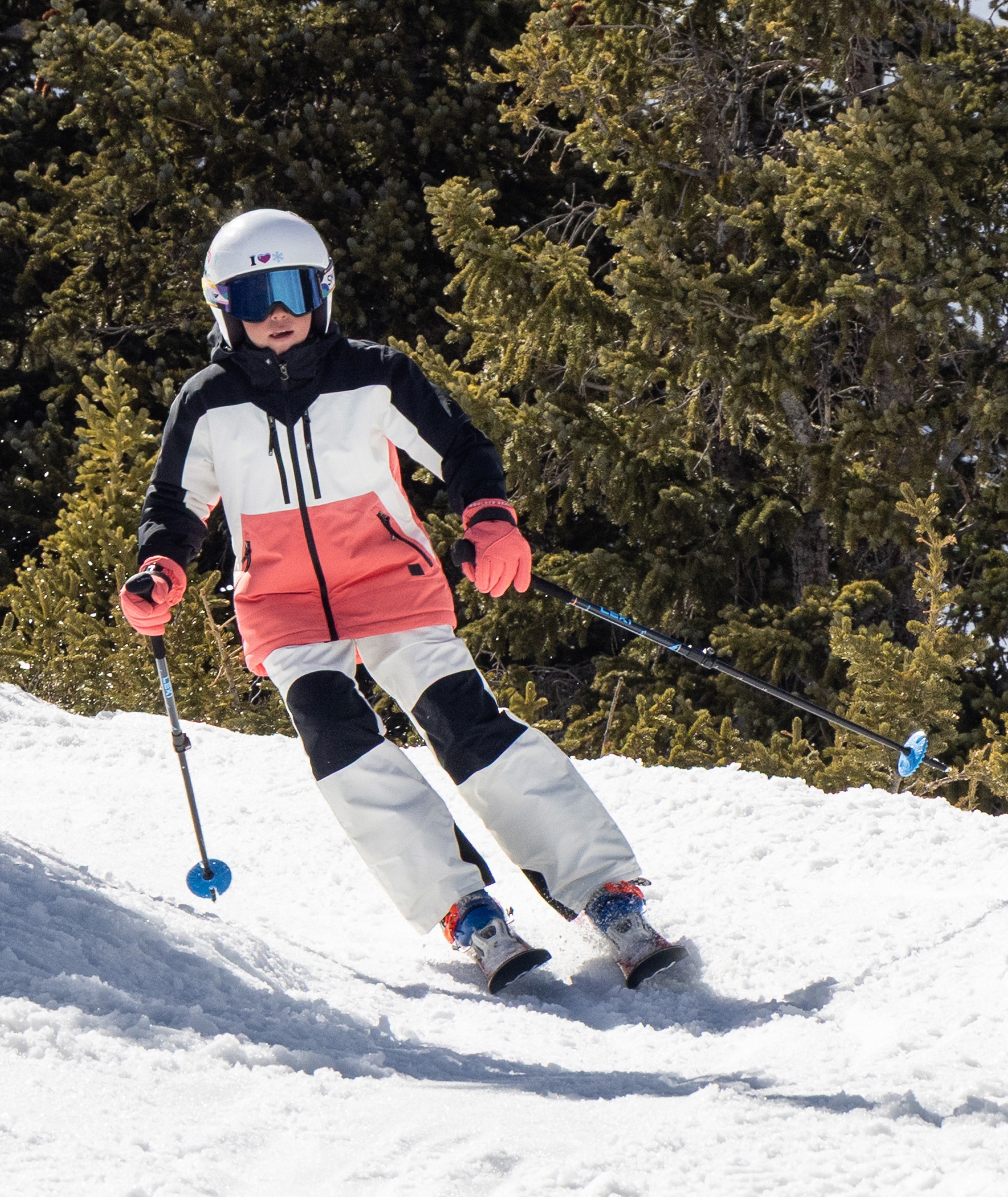 young person shown skiing down a snowy trail surrounded by pine trees shown wearing a black, white, and coral jacket with white pants.