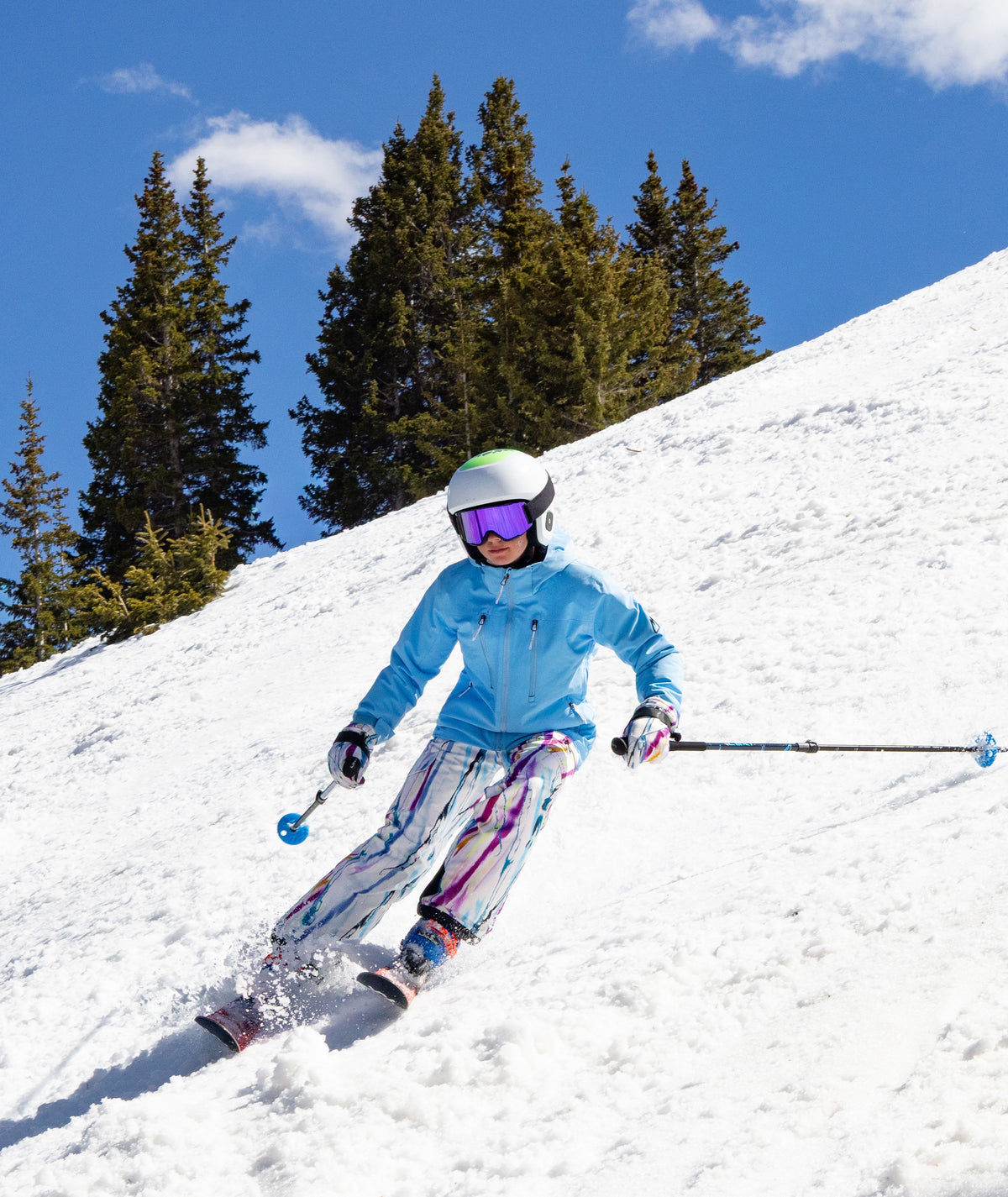 girl shown skiing down a snowy mountain in a light blue jacket with abstract multi color print pants. a blue sky and pine trees can be seen in the background.