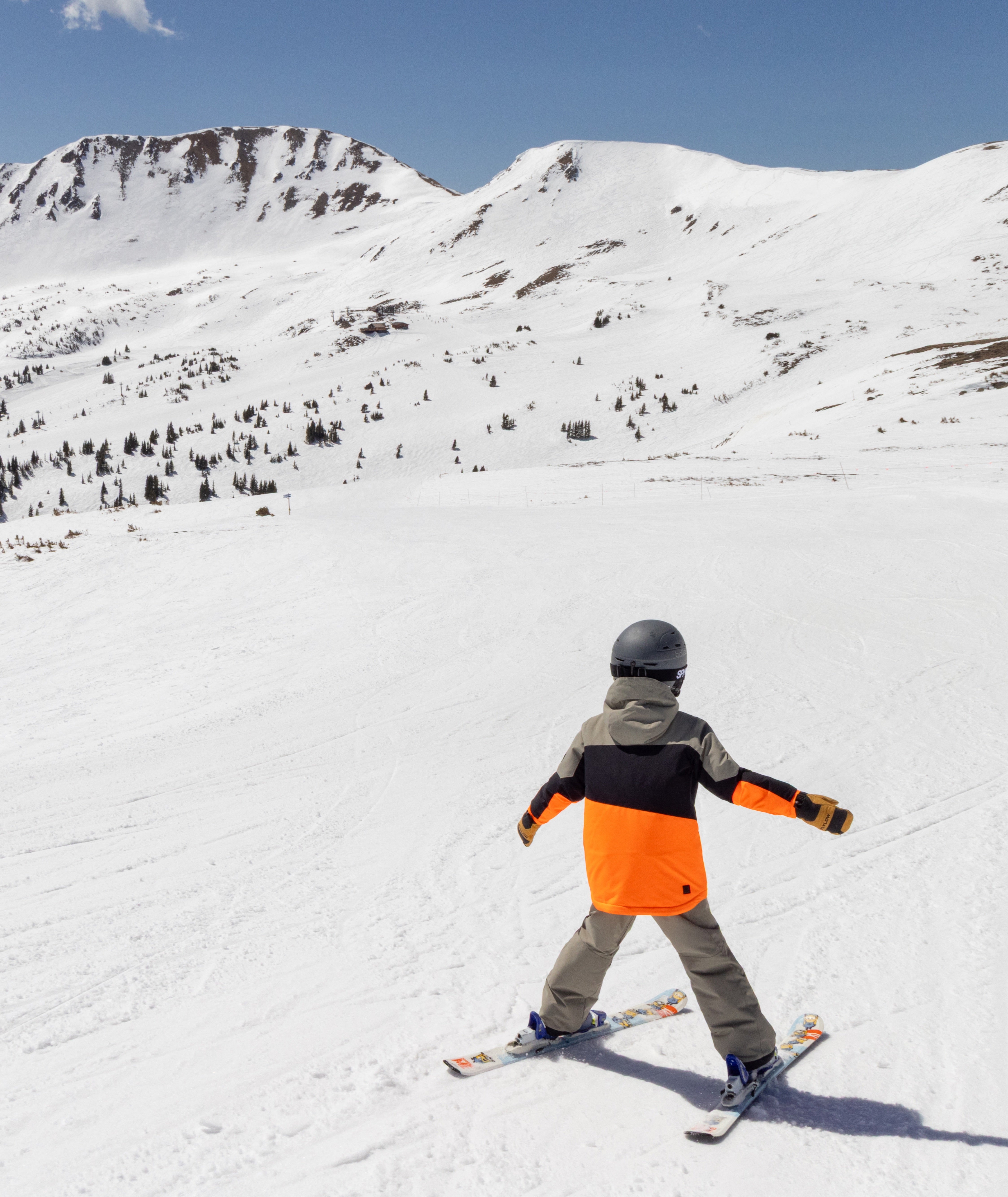 Person wearing an orange, beige and black jacket skiing on a snow-covered mountain with a clear blue sky