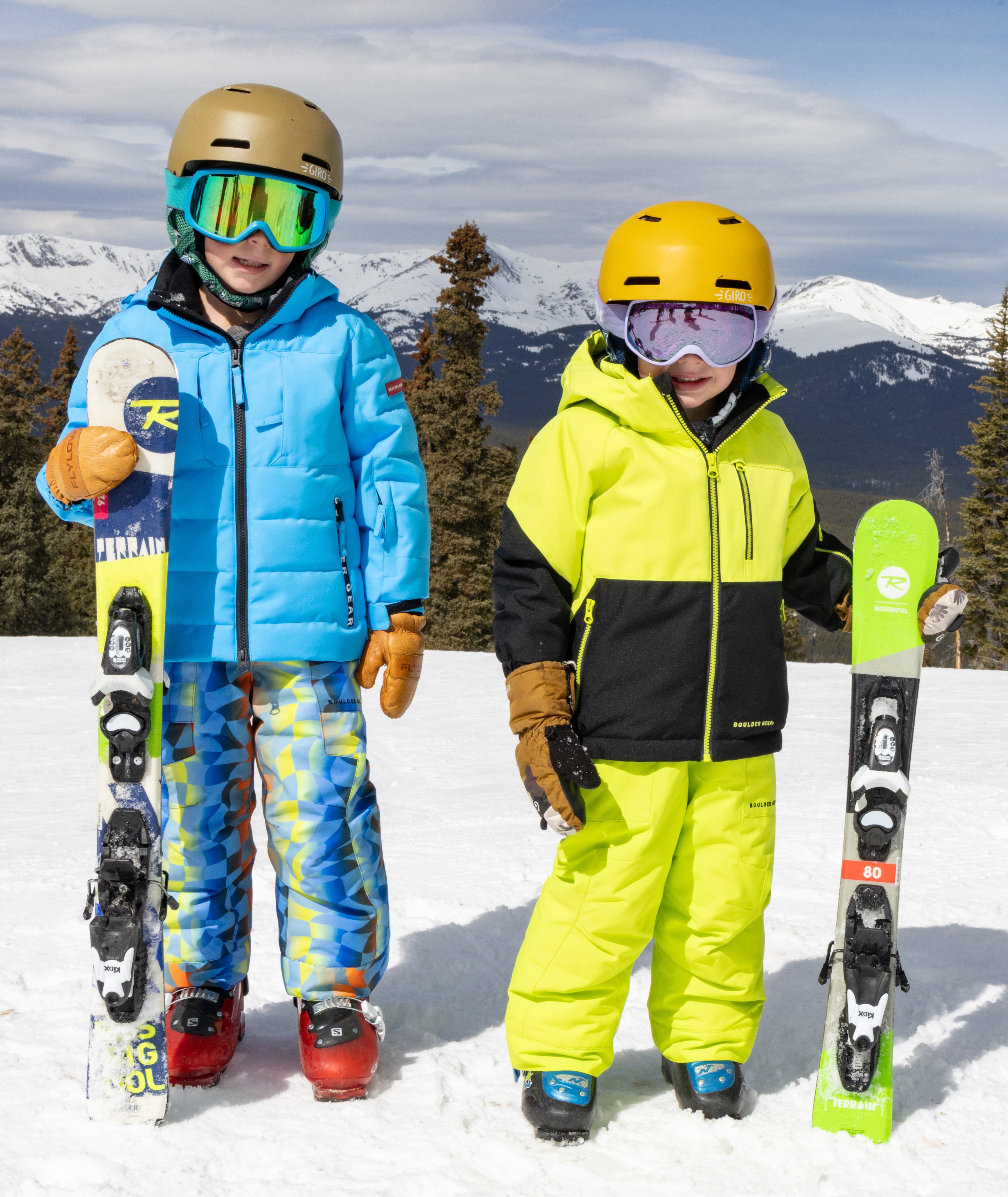 Two children in ski gear standing on a snowy slope with mountains in the background.
