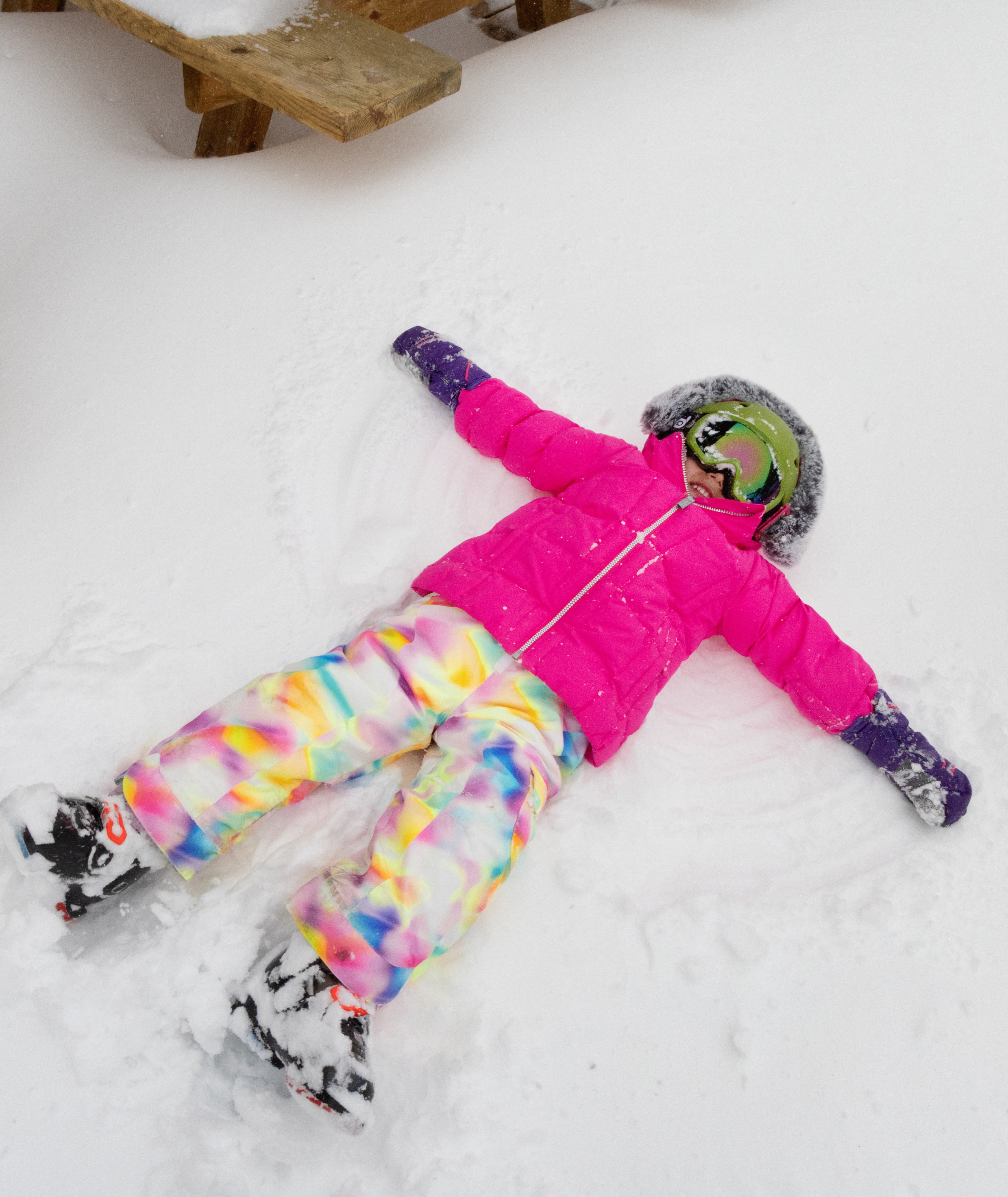 Person wearing a bright pink snowsuit with colorful pants lying in the snow.