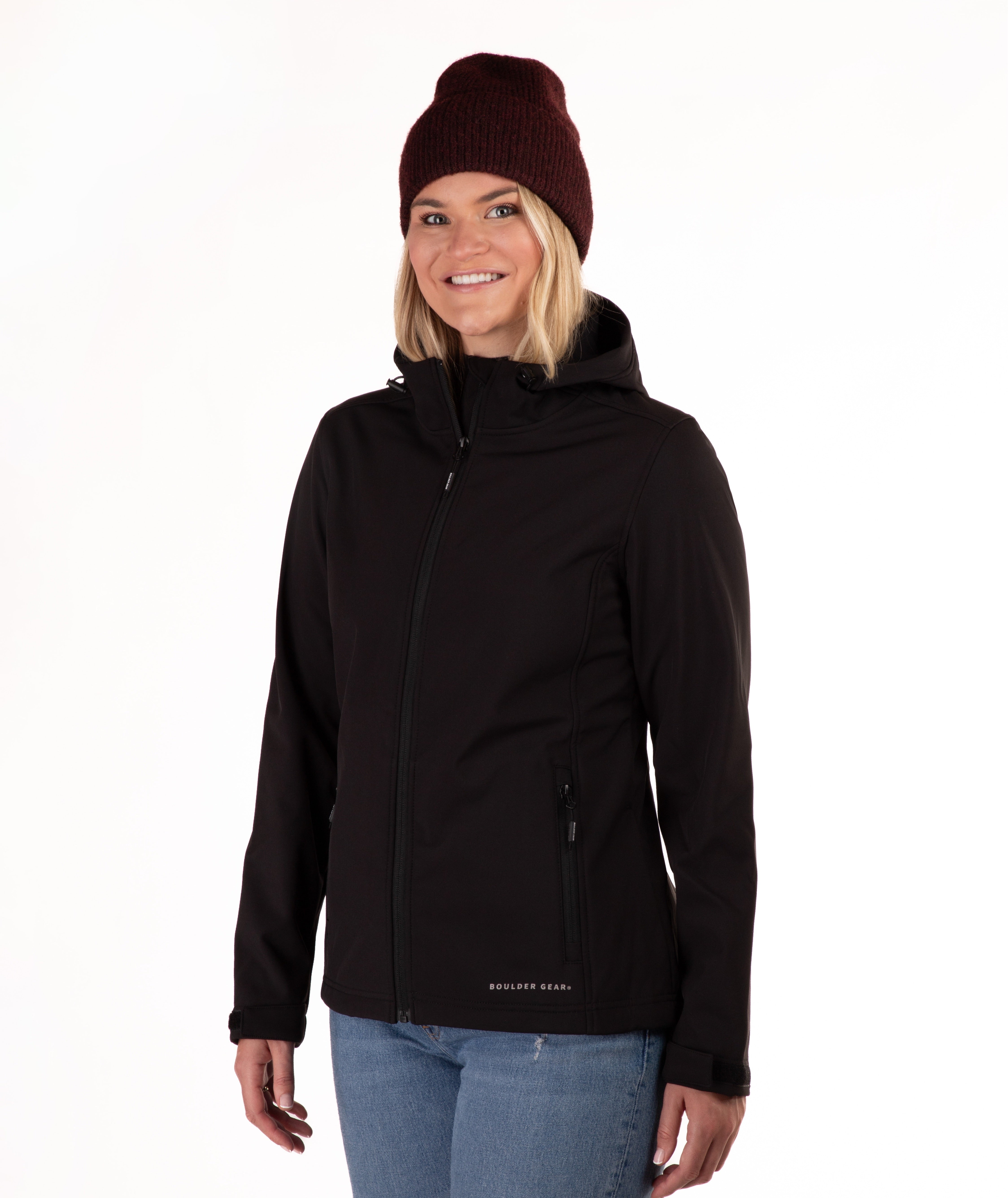 A woman wearing a black hooded jacket, a dark burgundy knit beanie, and blue jeans, standing against a plain white studio background.