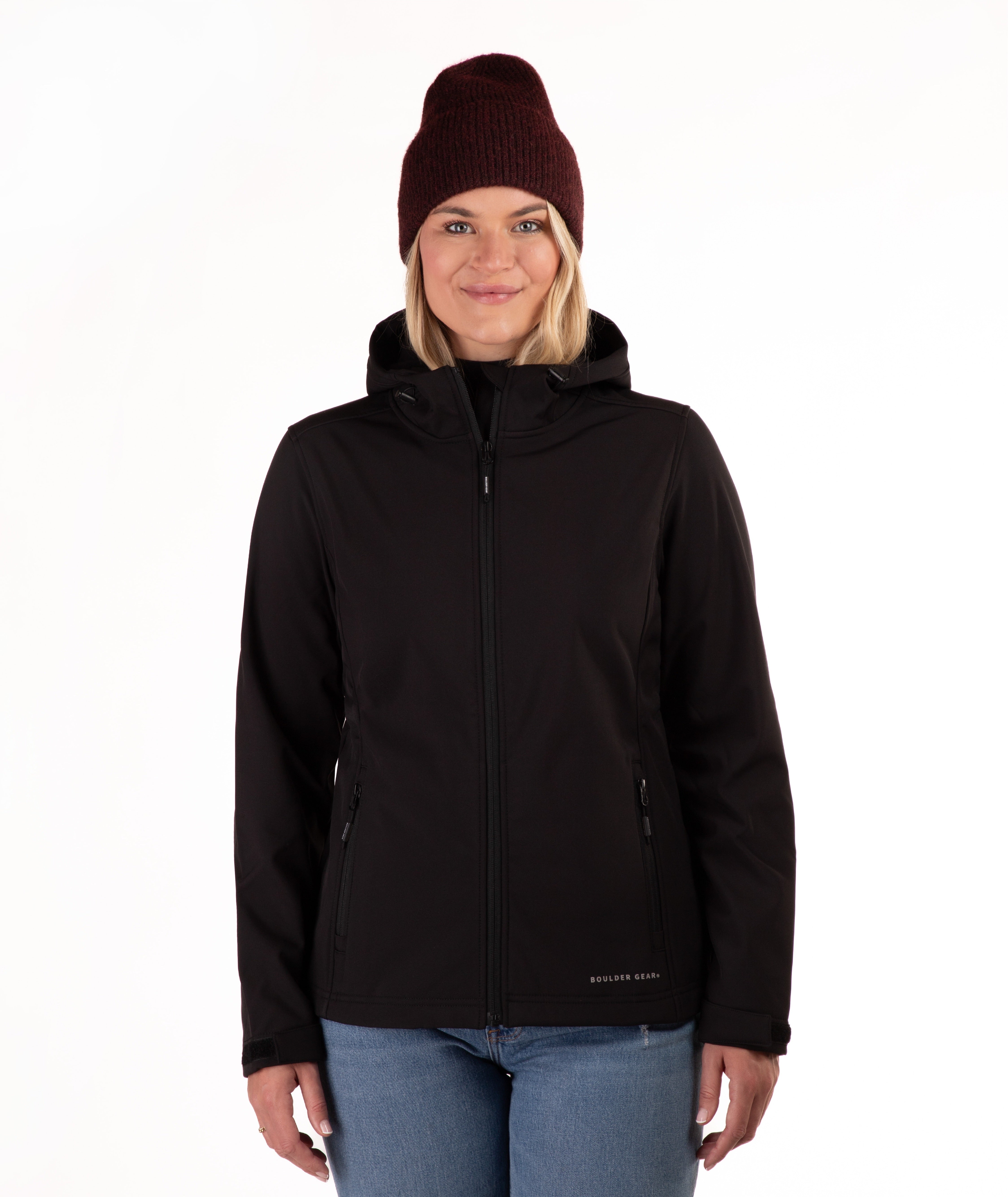 A woman wearing a black hooded zip-up jacket, blue jeans, and a dark burgundy knit beanie, standing against a plain white background.