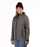 A woman wearing a gray hooded softshell jacket with black zippers, a dark burgundy knit beanie, and blue jeans, photographed against a plain white studio background.