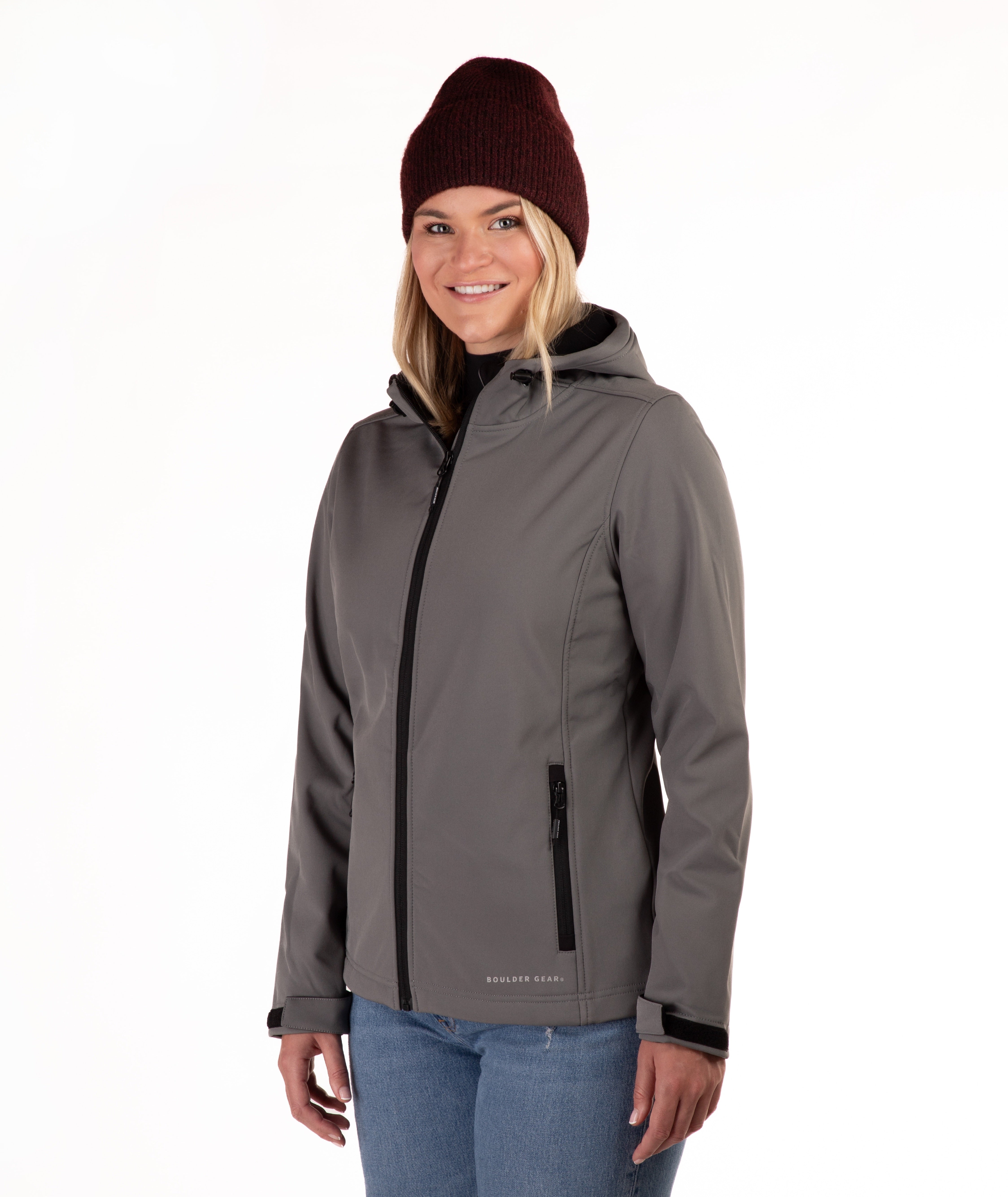 A woman wearing a gray hooded softshell jacket with black zippers, a dark burgundy knit beanie, and blue jeans, photographed against a plain white studio background.