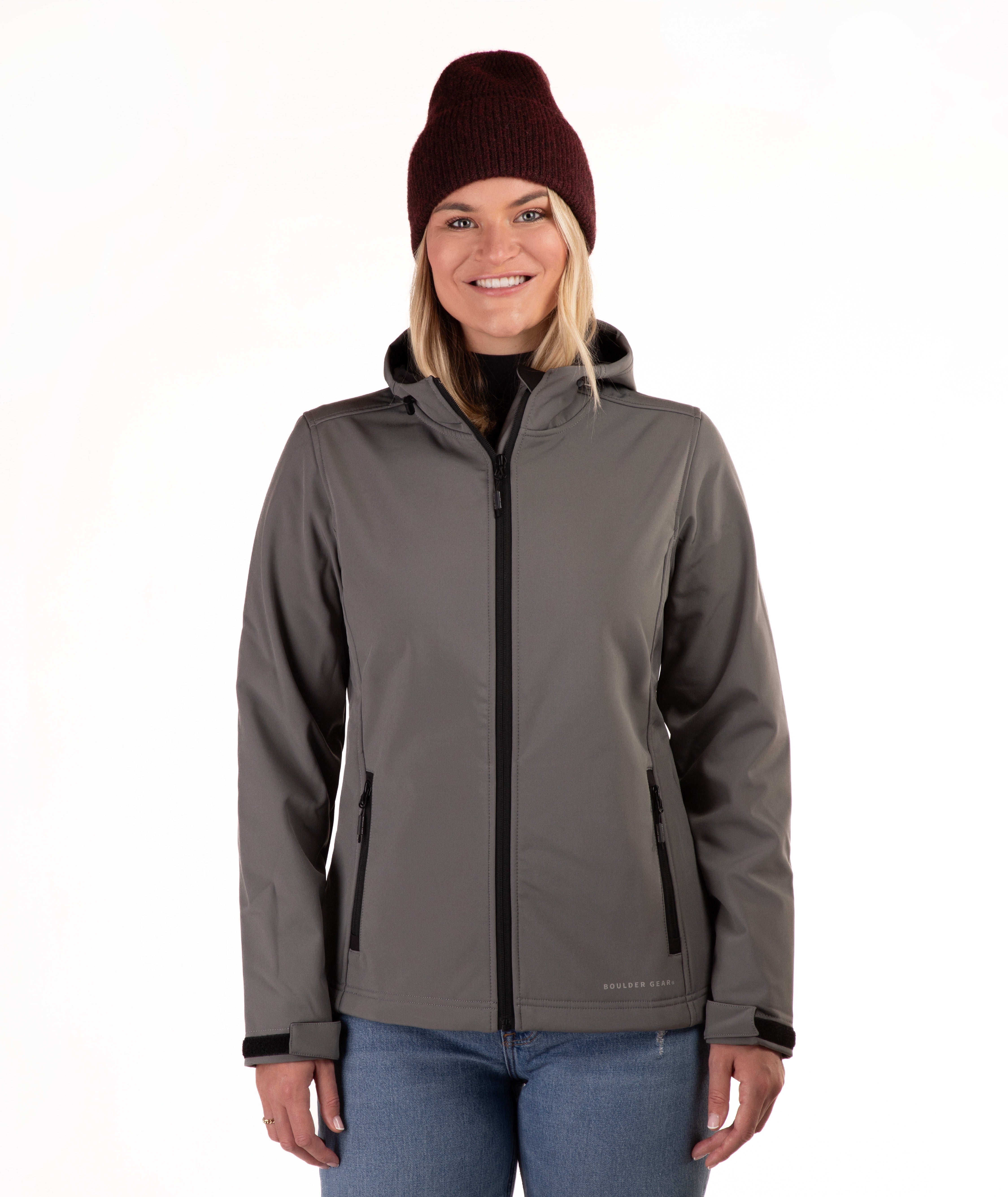A woman wearing a gray hooded zip-up jacket, a dark red knit beanie, and blue jeans, standing against a plain white background.