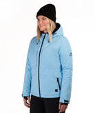 A woman wearing a light blue ski jacket with black zippers, a black beanie, and black pants, photographed against a plain white studio background.