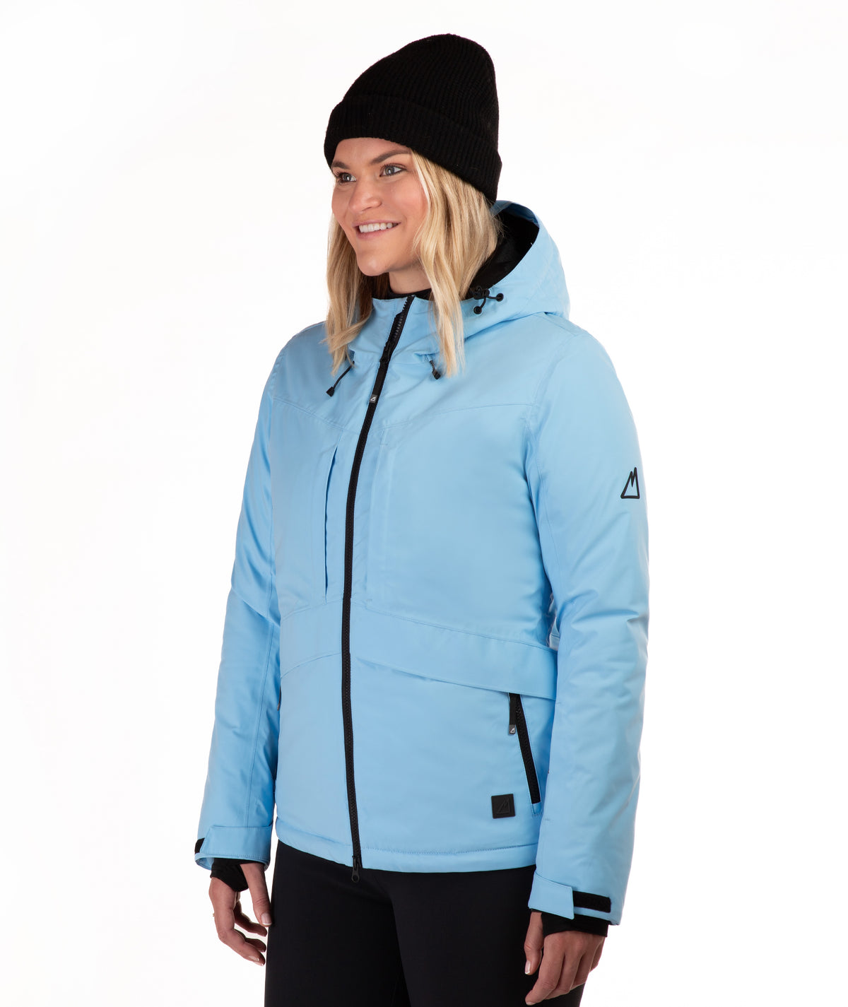 A woman wearing a light blue ski jacket with black zippers, a black beanie, and black pants, photographed against a plain white studio background.