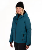 A woman wearing a teal ski jacket with black zippers, a black beanie, and black pants, photographed against a plain white studio background.