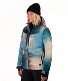A woman wearing a hooded technical ski jacket with a blue, beige, and rust-colored abstract print, paired with a black beanie, photographed against a solid white studio background.