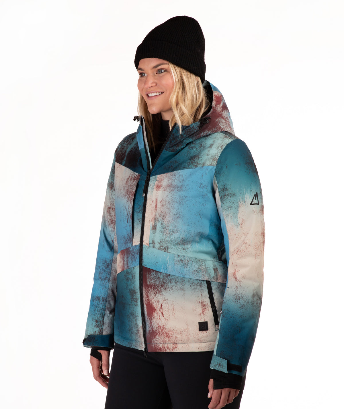 A woman wearing a hooded technical ski jacket with a blue, beige, and rust-colored abstract print, paired with a black beanie, photographed against a solid white studio background.