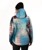 A person shown from the back wearing a hooded jacket with an abstract watercolor-style print in blue, teal, beige, and muted reddish-brown tones, paired with a black beanie and black pants against a plain white studio background.