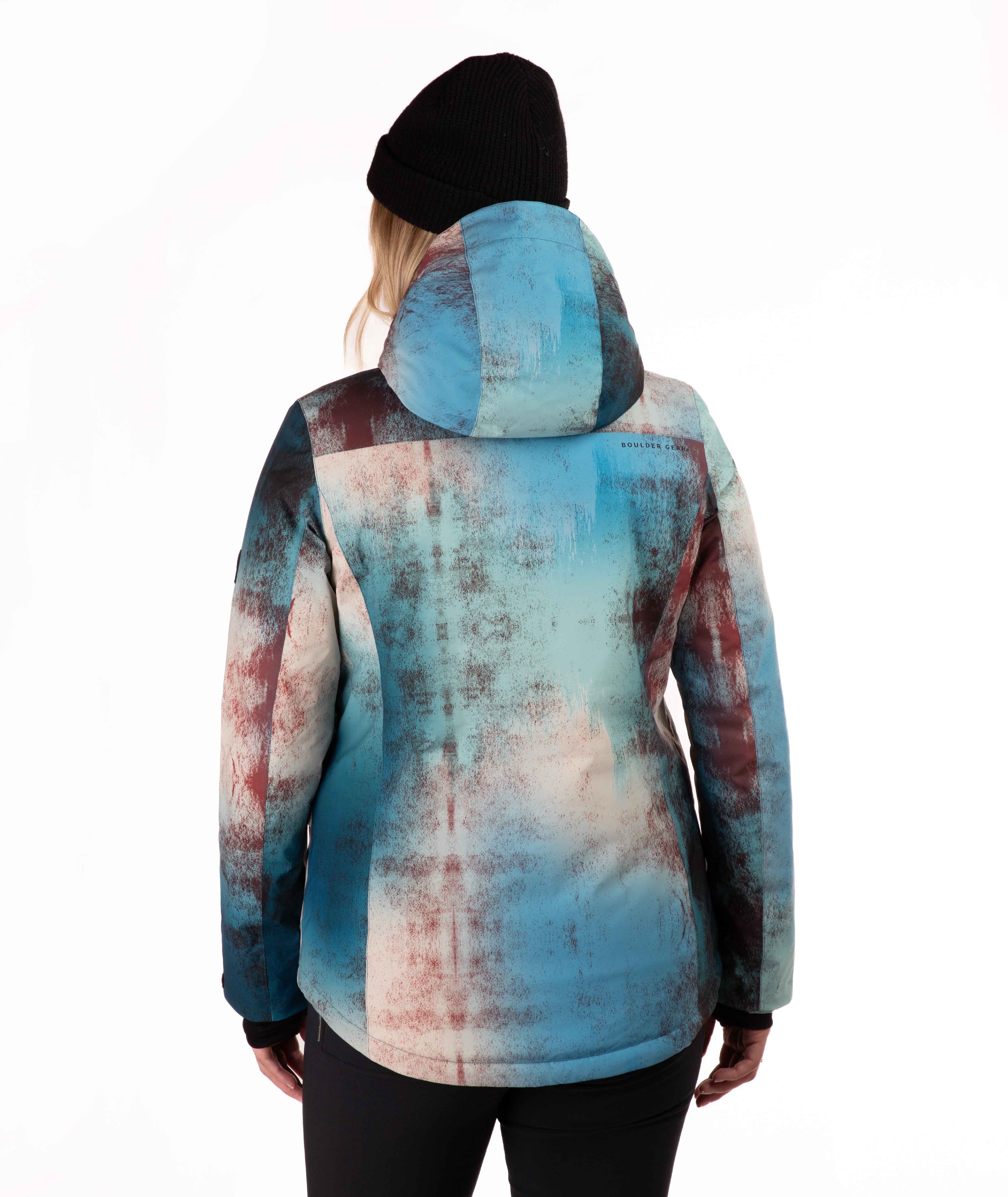 A person shown from the back wearing a hooded jacket with an abstract watercolor-style print in blue, teal, beige, and muted reddish-brown tones, paired with a black beanie and black pants against a plain white studio background.