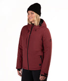 A woman wearing a burgundy hooded ski jacket with black zippers, a black beanie, and black pants, photographed against a plain white studio background.