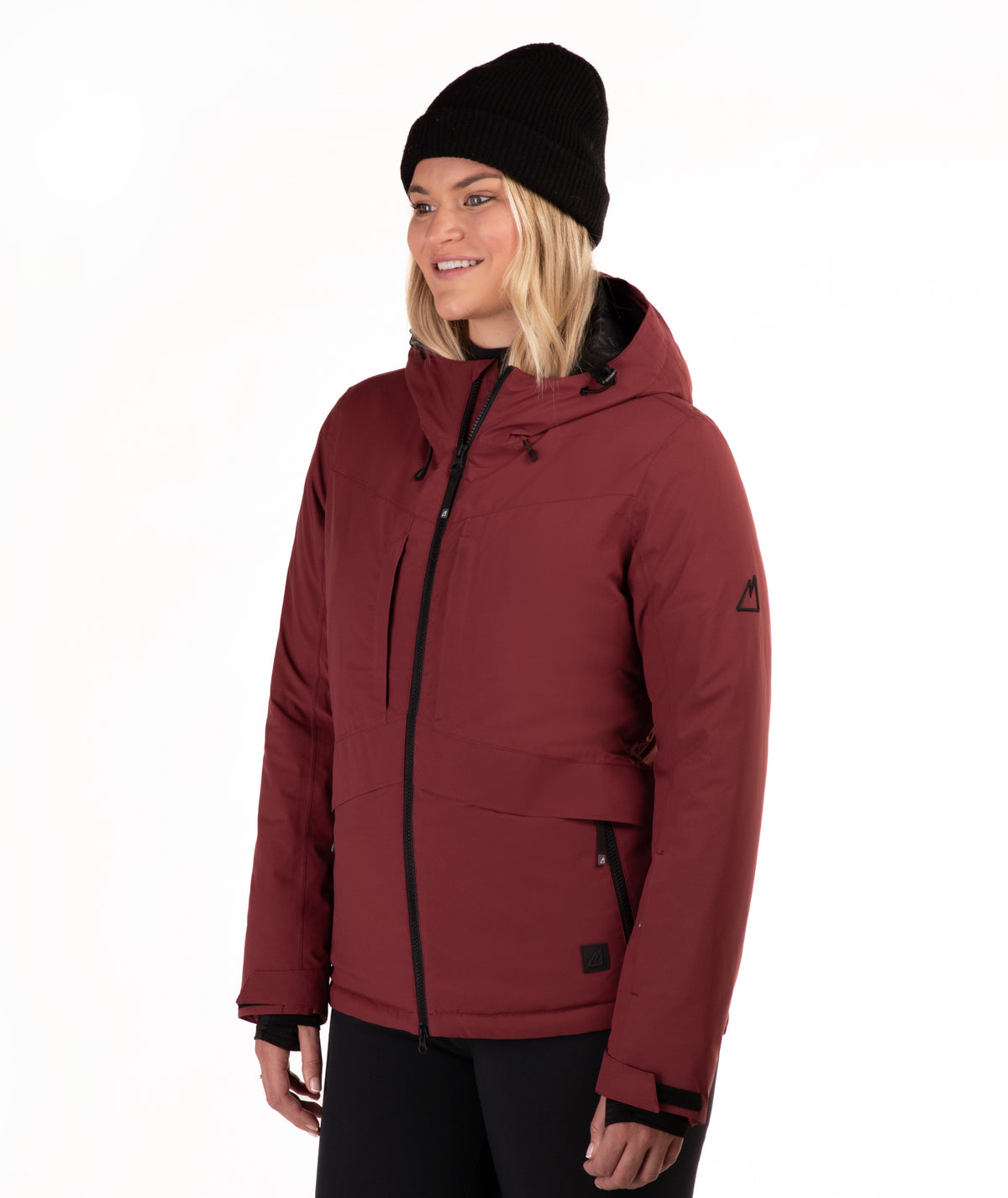 A woman wearing a burgundy hooded ski jacket with black zippers, a black beanie, and black pants, photographed against a plain white studio background.