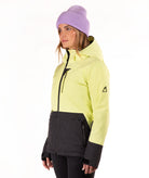 A woman wearing a light yellow ski jacket with a black patterned lower panel and black zipper details, paired with a light purple knit beanie, shown in profile against a white studio background.