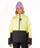 A woman wearing a light yellow and black ski jacket with a purple knit beanie, standing against a plain white background.