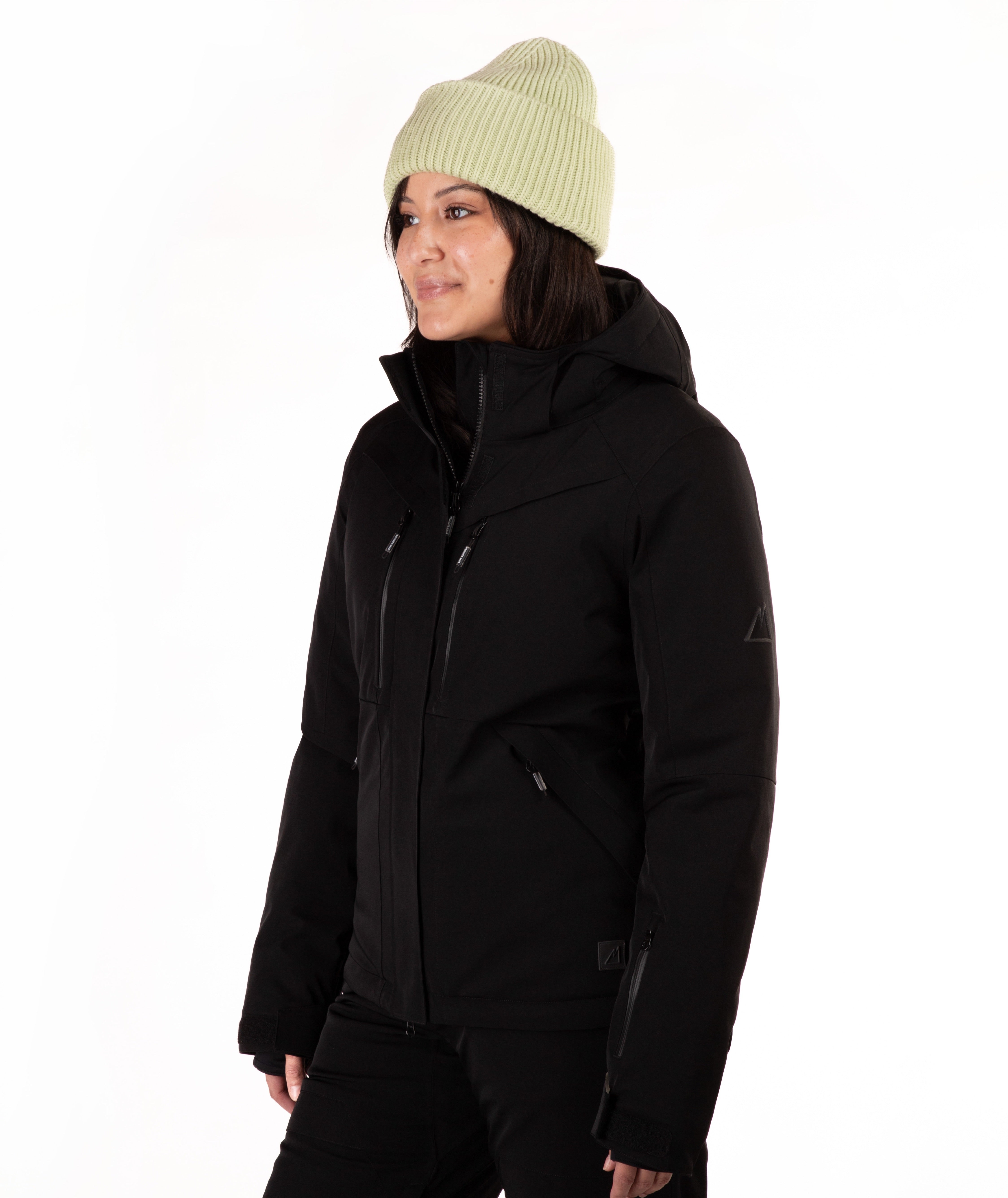 A woman wearing a black ski jacket and matching black pants, paired with a light green knit beanie, standing against a plain white studio background.