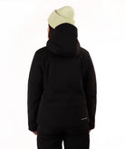 A woman wearing a black ski jacket and matching black pants, paired with a light green knit beanie, standing against a plain white studio background.