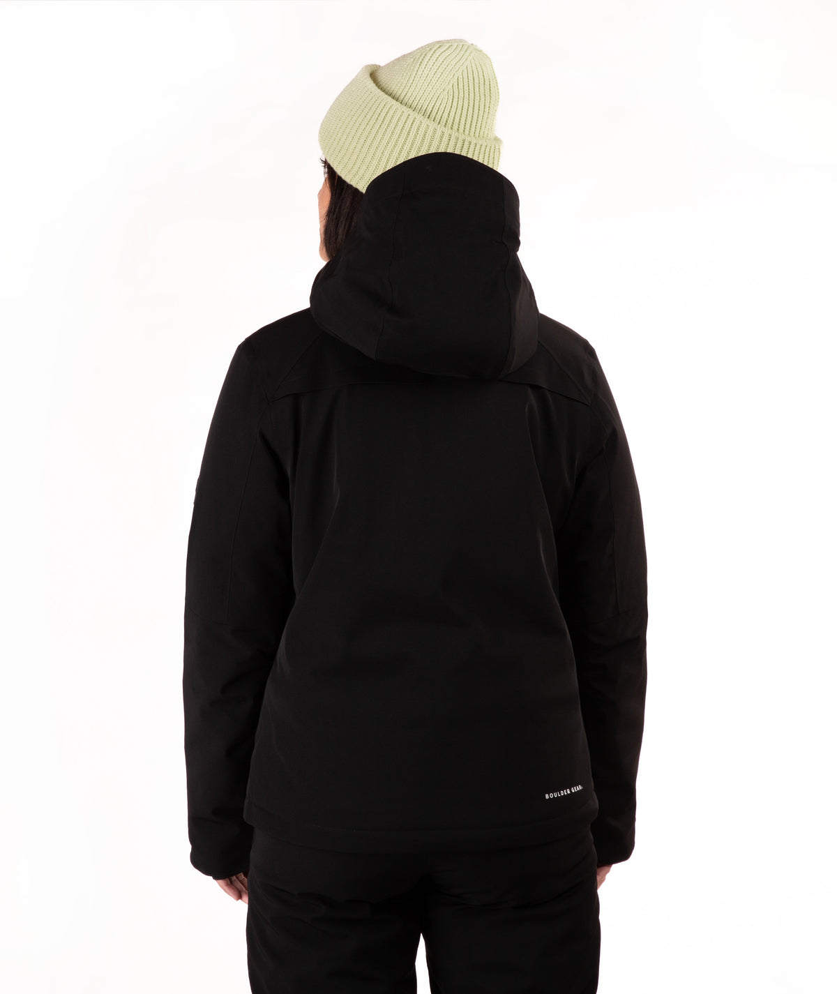 A woman wearing a black ski jacket and matching black pants, paired with a light green knit beanie, standing against a plain white studio background.