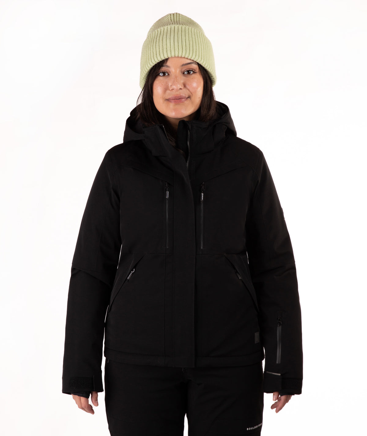 A woman wearing a black ski jacket and matching black pants, paired with a light green knit beanie, standing against a plain white studio background.