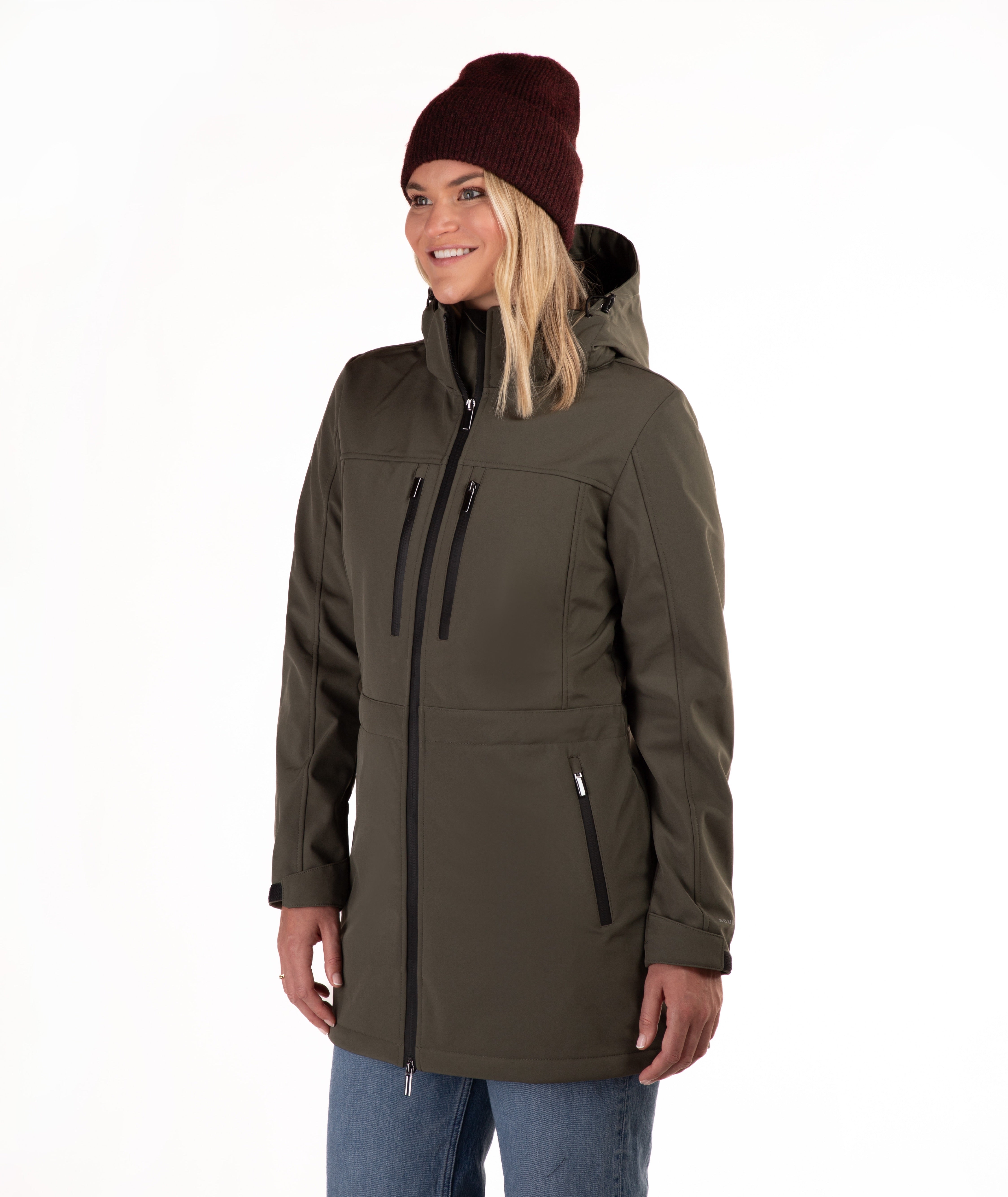 A woman wearing an olive green hooded jacket with black zippers, blue jeans, and a dark red knit beanie, photographed against a plain white studio background.