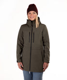 A woman wearing a dark olive green hooded softshell jacket with black zippers, blue jeans, and a burgundy knit beanie, standing against a plain white background.