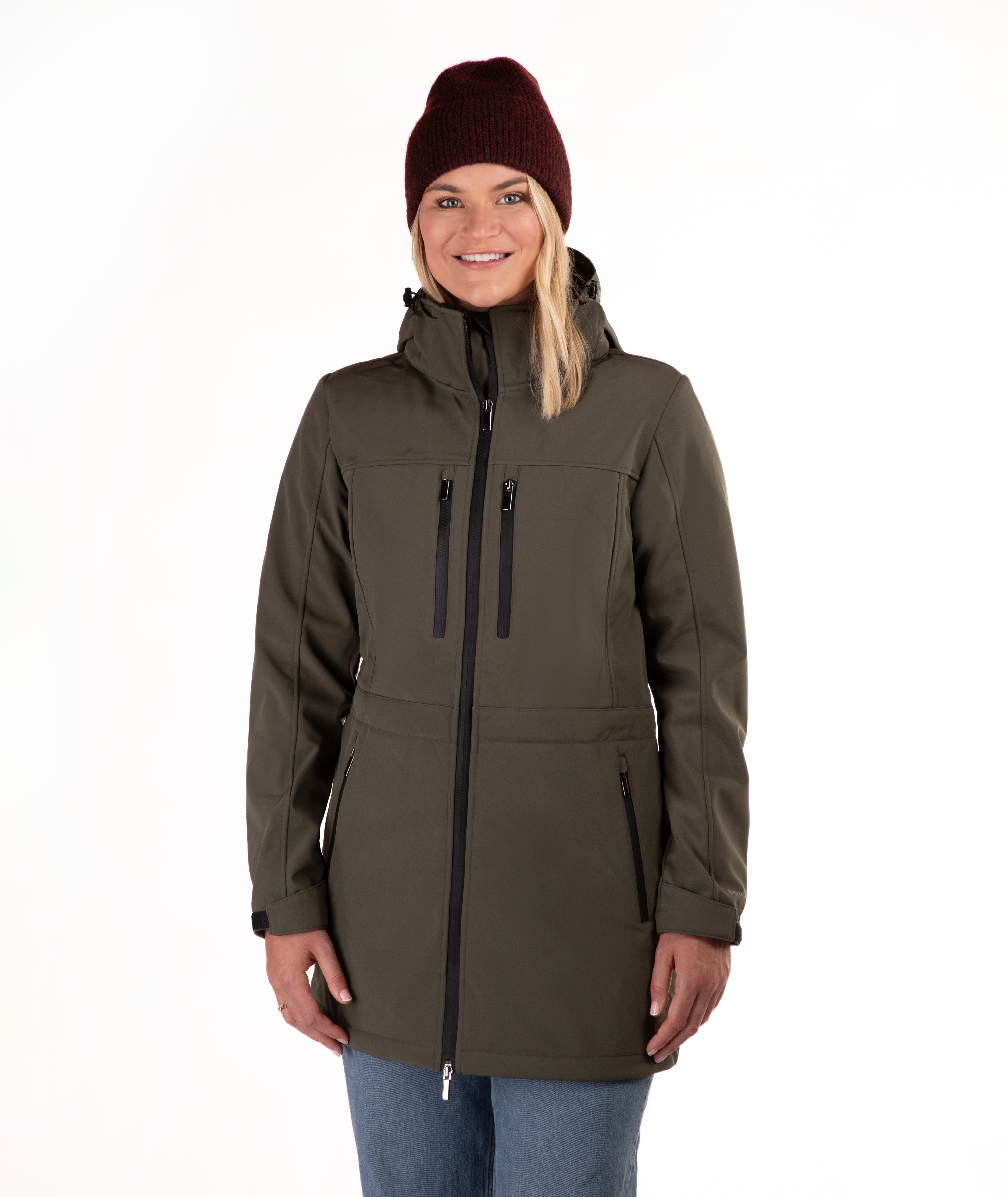 A woman wearing a dark olive green hooded softshell jacket with black zippers, blue jeans, and a burgundy knit beanie, standing against a plain white background.