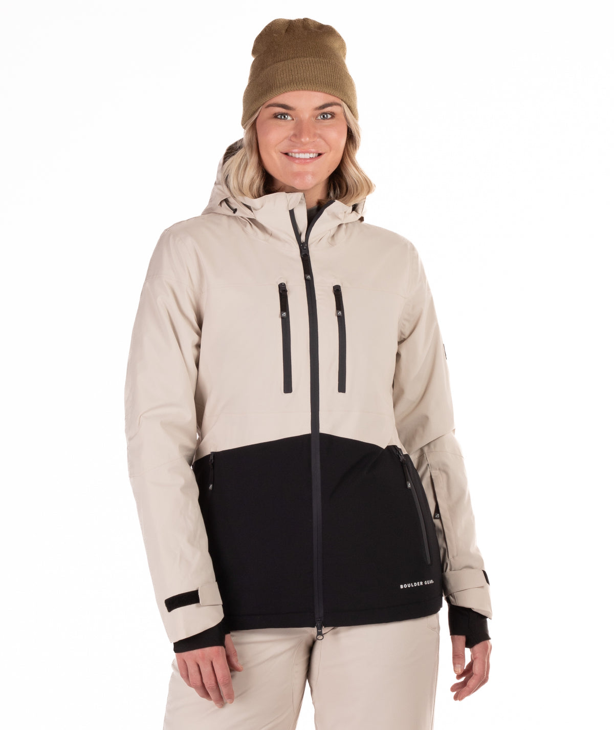 A woman wearing a beige and black snowboard jacket, a tan beanie, and beige snow pants, standing against a solid white background.