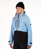 A woman wearing a light blue and black color-blocked ski jacket, a black beanie, and black pants, photographed against a plain white studio background.