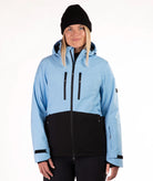A woman wearing a light blue and black color-blocked ski jacket, a black beanie, and black pants, photographed against a plain white studio background.