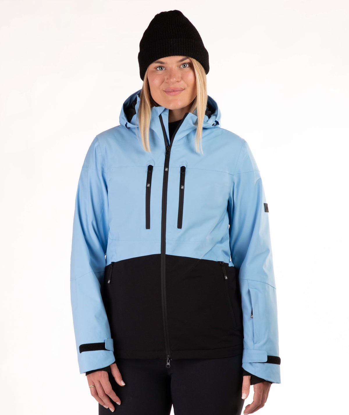 A woman wearing a light blue and black color-blocked ski jacket, a black beanie, and black pants, photographed against a plain white studio background.