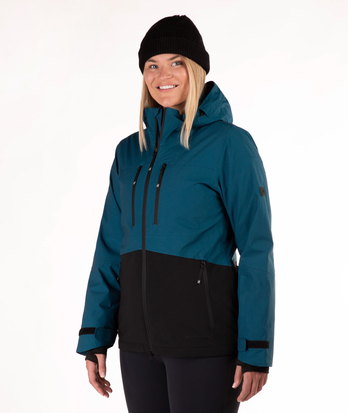 A woman wearing a dark teal and black color-blocked ski jacket, a black beanie, and black pants, standing against a plain white studio background.
