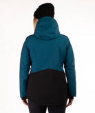 A woman wearing a dark teal and black color-blocked ski jacket, a black beanie, and black pants, standing against a plain white studio background.